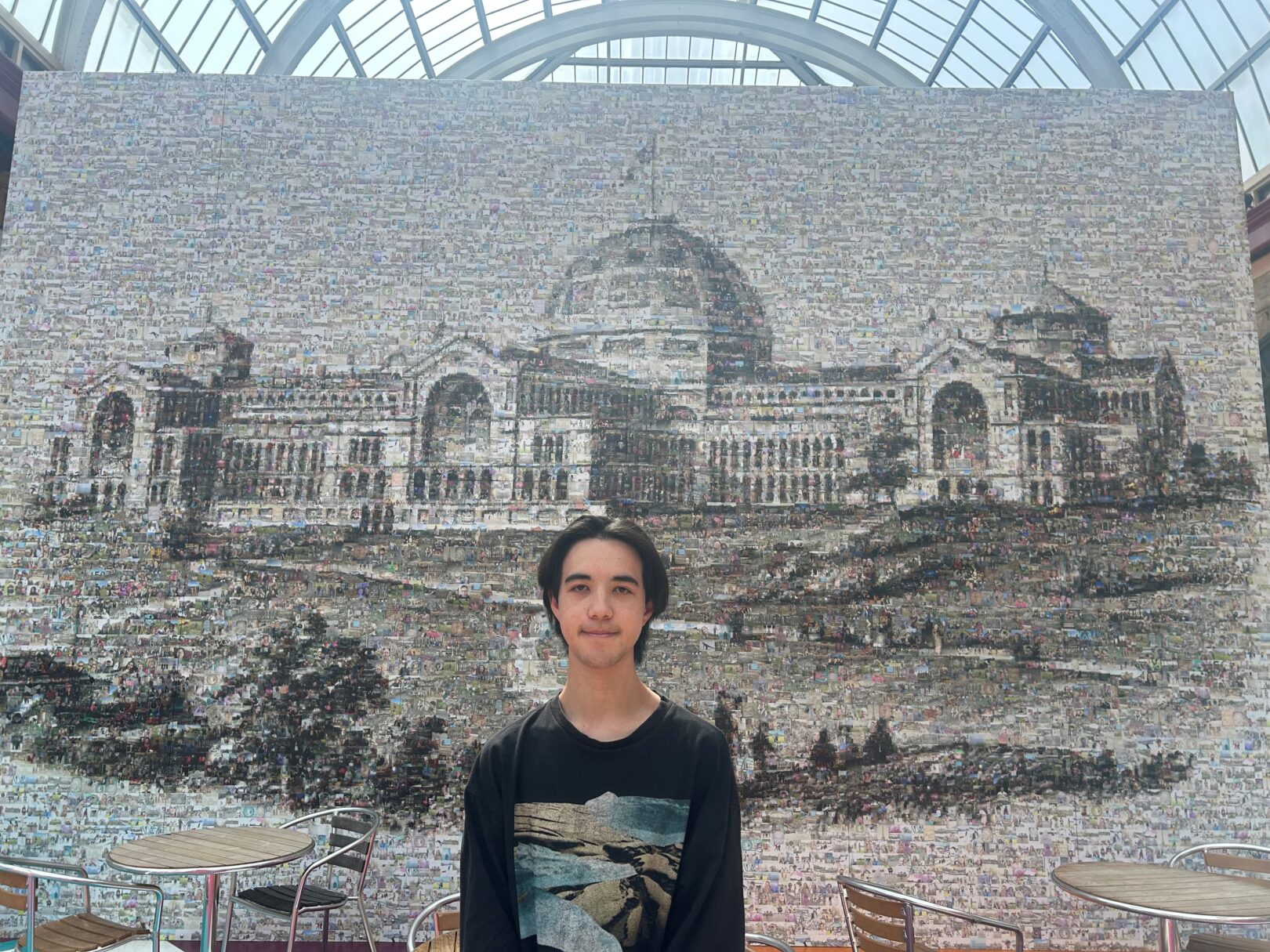 A young man wearing a dark long sleeve top stands in a large room with a glass roof, in front of mural of Alexandra Palace, London.