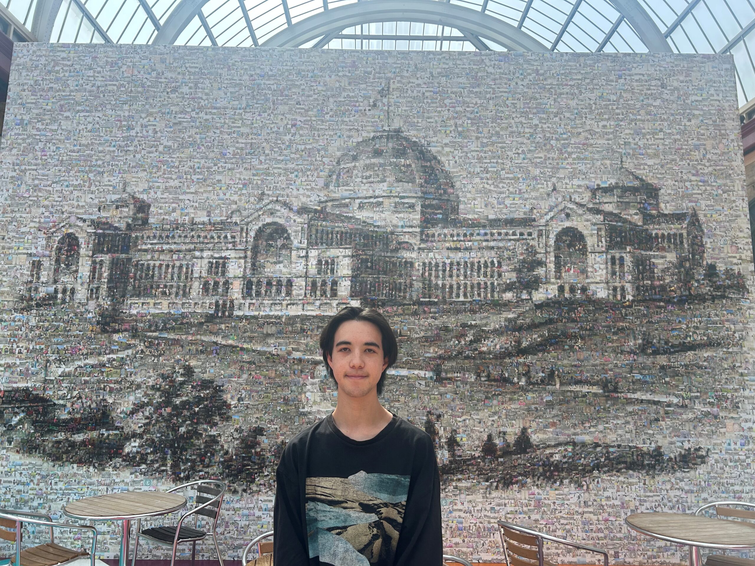 A young man wearing a dark long sleeve top stands in a large room with a glass roof, in front of mural of Alexandra Palace, London.