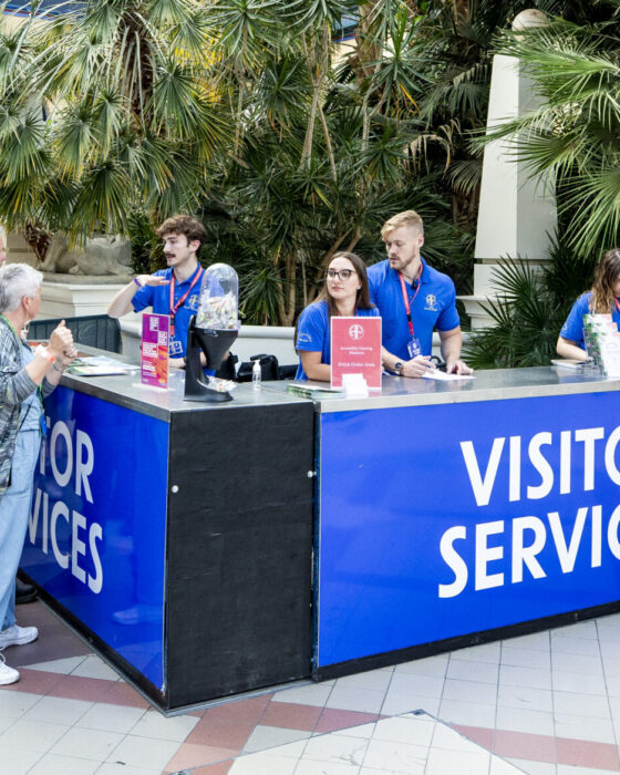 A visitor services desk in a bright, spacious atrium with large plants. Staff in blue shirts assist visitors. A line forms, suggesting a busy, welcoming atmosphere.