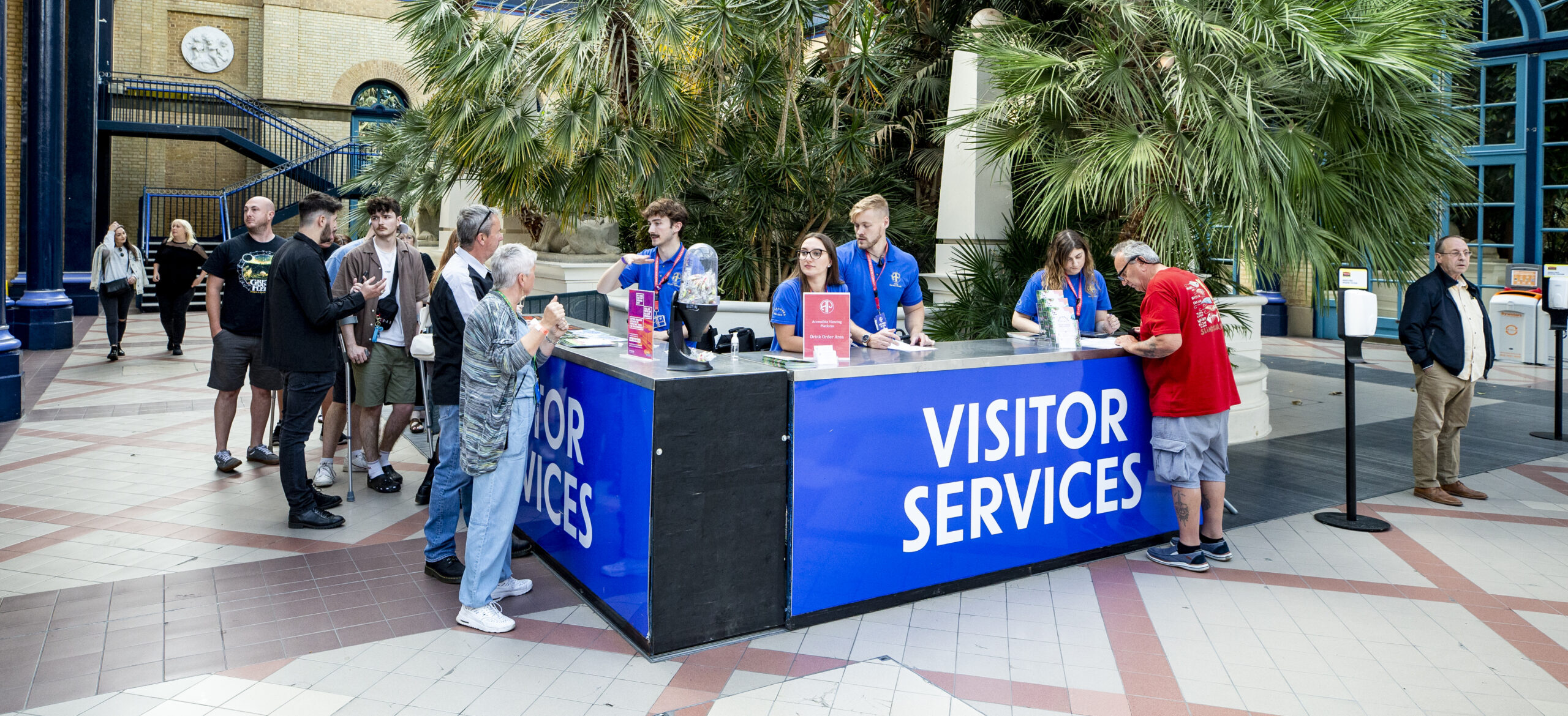 A visitor services desk in a bright, spacious atrium with large plants. Staff in blue shirts assist visitors. A line forms, suggesting a busy, welcoming atmosphere.